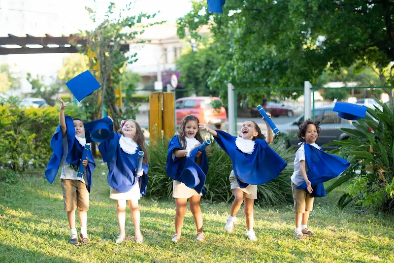 Crianças felizes com toga em formatura infantil jogando quepe para cima